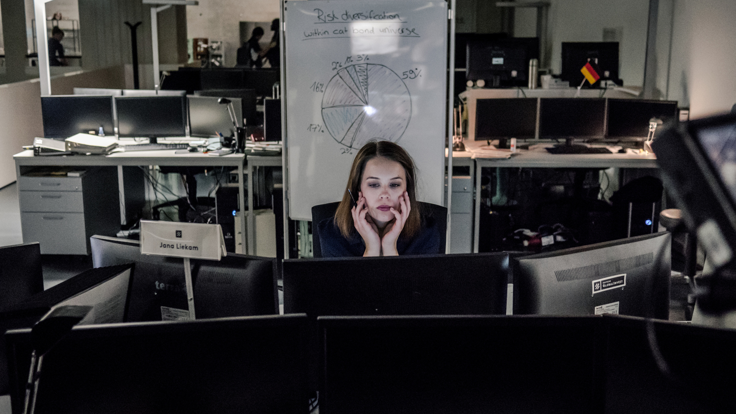 Jana Liekam (Paula Beer) sitzt an ihrem Schreibtisch hinter mehreren Bildschirmen und arbeitet. Es scheint spät zu sein, denn die anderen Arbeitsplätze im Großraumbüro sind leer.
