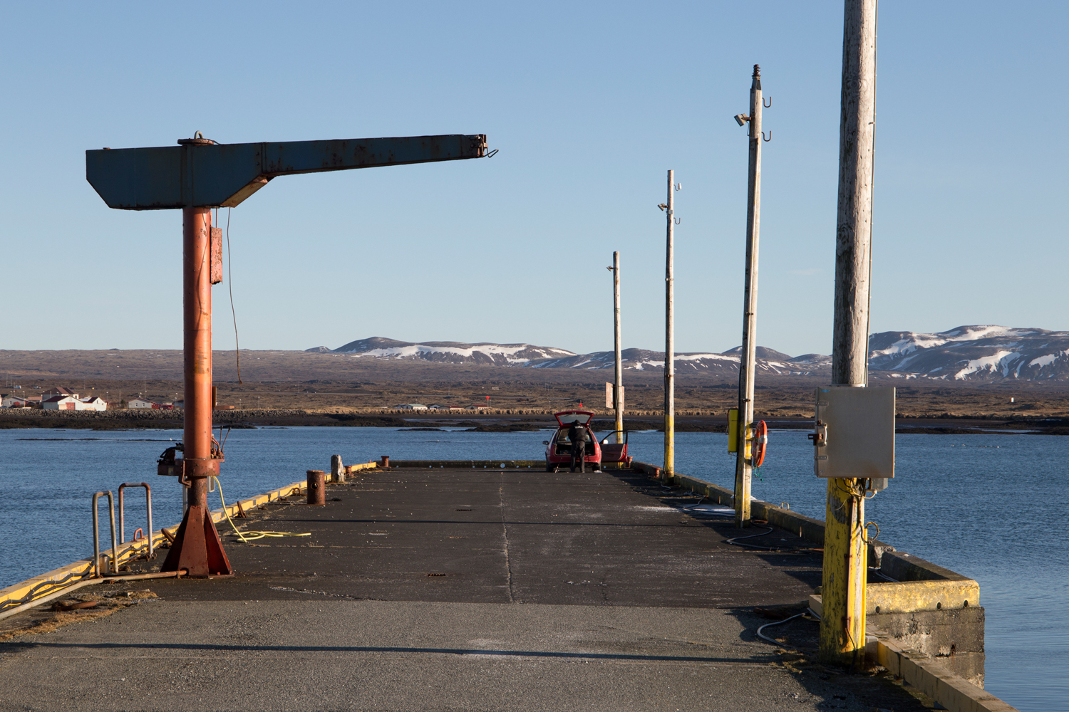 A man has parked his tiny red car at the end of a pier and is unloading his fishing gear while the sun is shining on a deserted harbour framed by distant mountains.