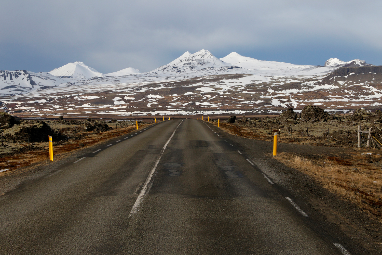 An empty street stretches from the foreground to the horizon in an almost straight line, leading through fields of volcanic rocks with distinctive green lichens growing on the surface towards a jagged mountain chain that is covered in snow.