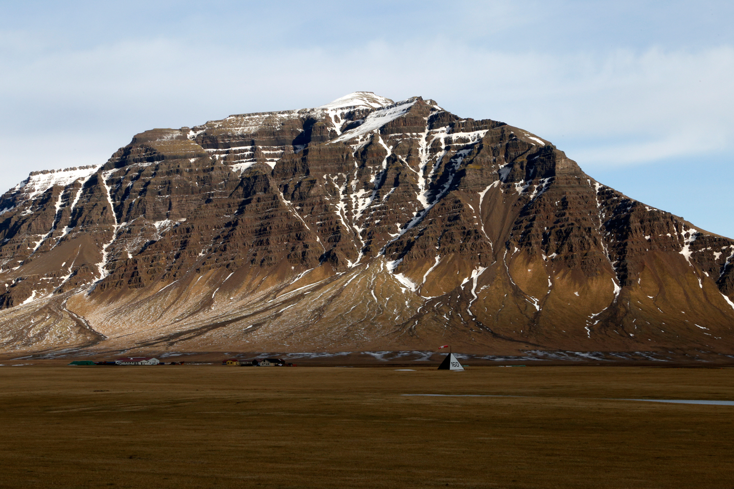 At the bottom of a volcanic mountain that was formed by layers of rock piling up on top of each other in a highly geometrical fashion, there is a small tent in the shape of a tetrahedron with a windsock fixed on top.