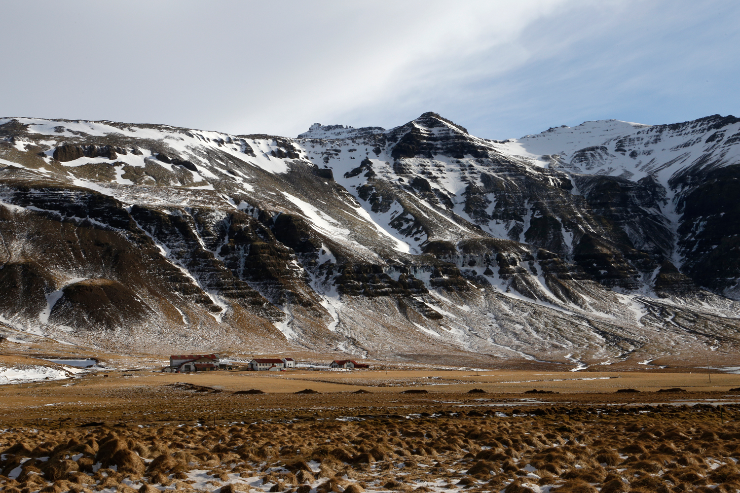 A farm at the bottom of a very steep mountain chain, sitting in a field of dry grass with some occasional spots of snow.