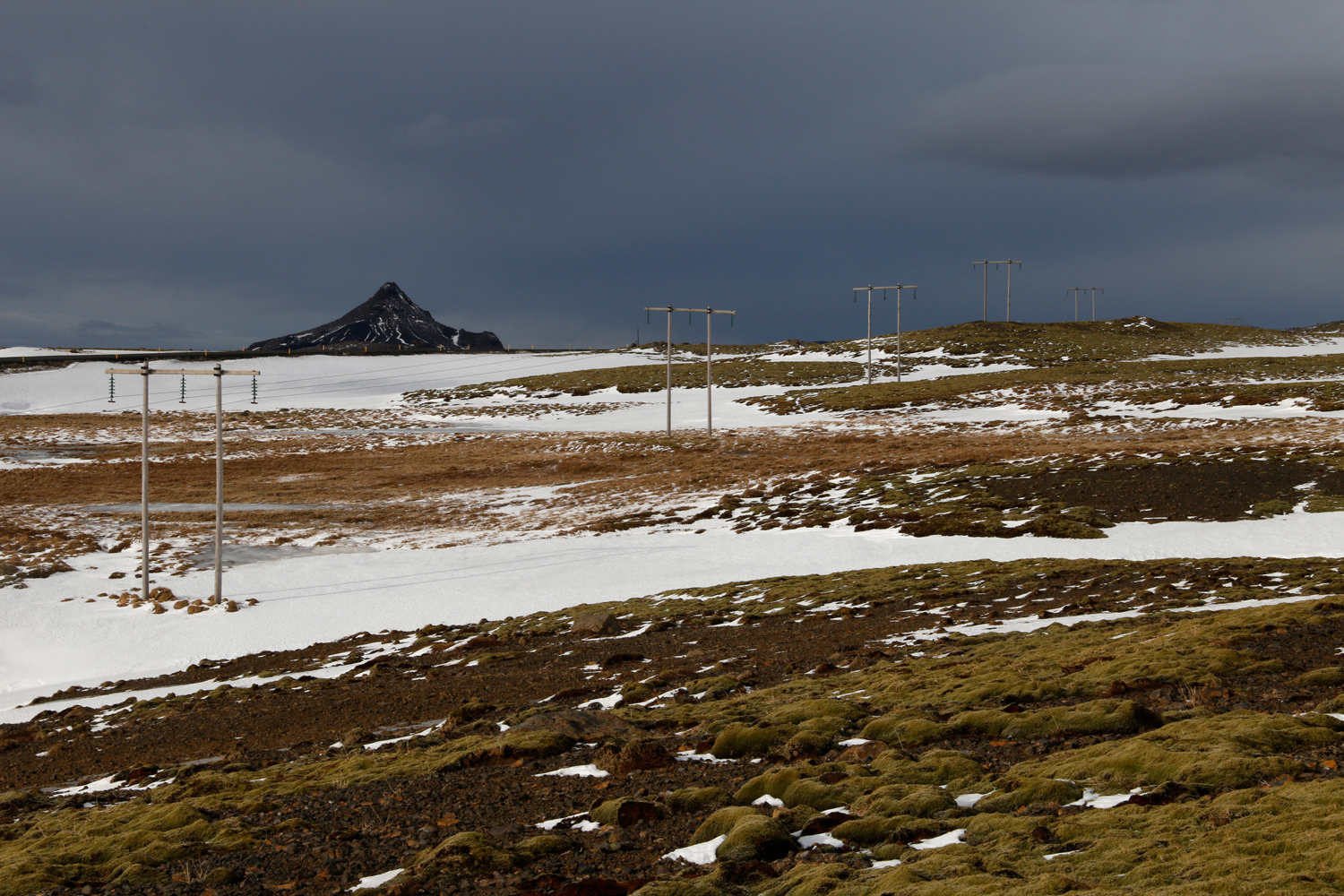 Transmission towers lead up a hill that is covered in the green of lichens, the white of unspoilt snow and the light brown of dry grass, with a black, jagged mountain in the distance and dark clouds looming above.