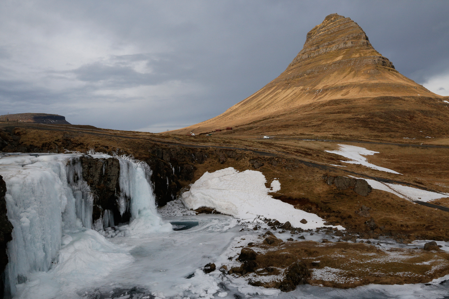 A view of Mount Kirkjufell and Kirkjufellsfoss waterfall, which is mostly frozen. It is a peaceful scene, although some faint rays of sunlight are contrasted with dark silver clouds.