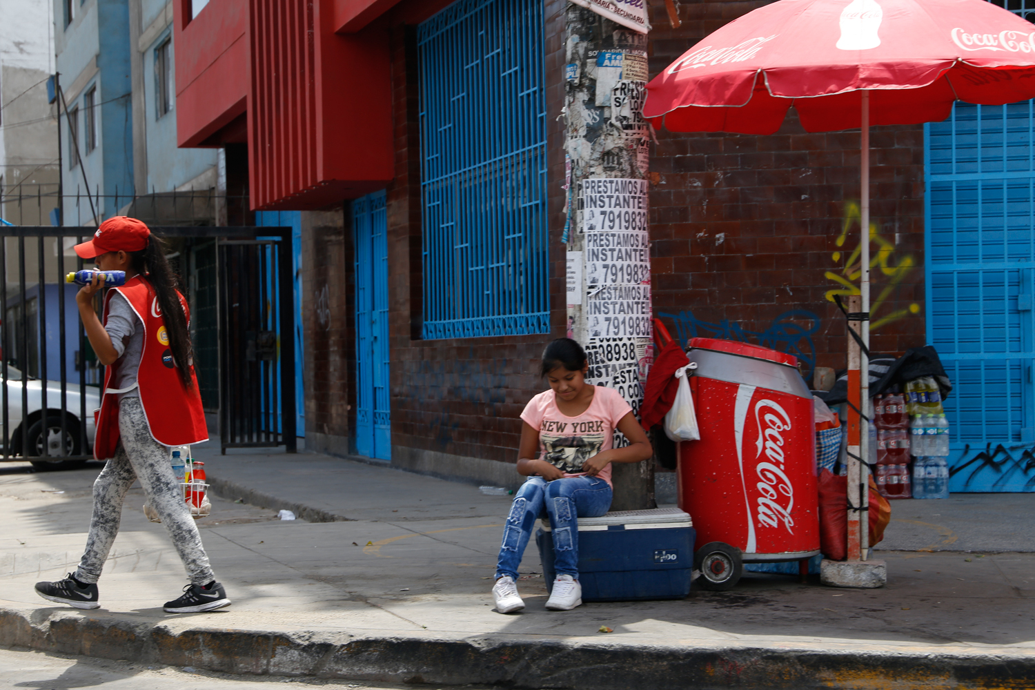 Two girls operate a mobile kiosk on the side of a road. One of them is sitting on a plastic box next to a stack of bottled lemonade, shaded by a red Coca Cola parasol. The other one is walking along the curb with a small crate of plastic bottles to be sold to people passing by in cars.