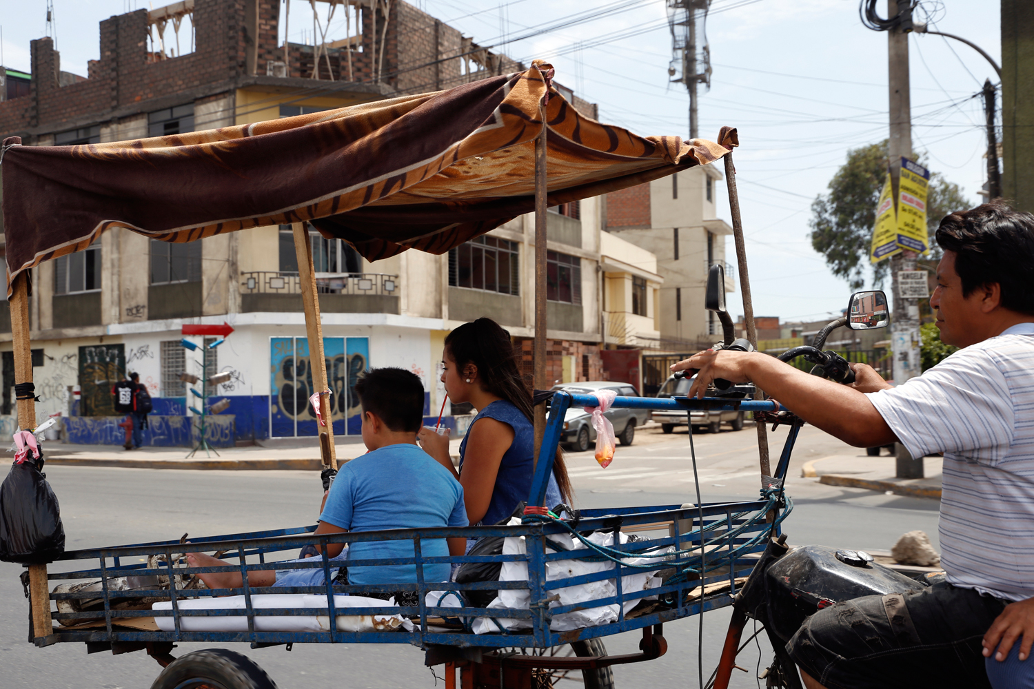 In what appears to be a family trip, a man rides a motorcycle with a two-wheeled cart attached to its front. There's a second person riding the motorbike and two children are sitting in the cart, drinking from plastic cups with red straws to refresh themselves on a sunny day.