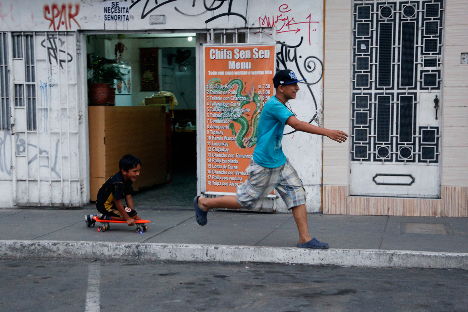 Two boys race along the pavement in front of a typical Chinese-Peruvian Chifa restaurant. The older one in his Crocs-style shoes is running from the younger one who is kneeling on a red plastic skateboard. Both are laughing heartily.