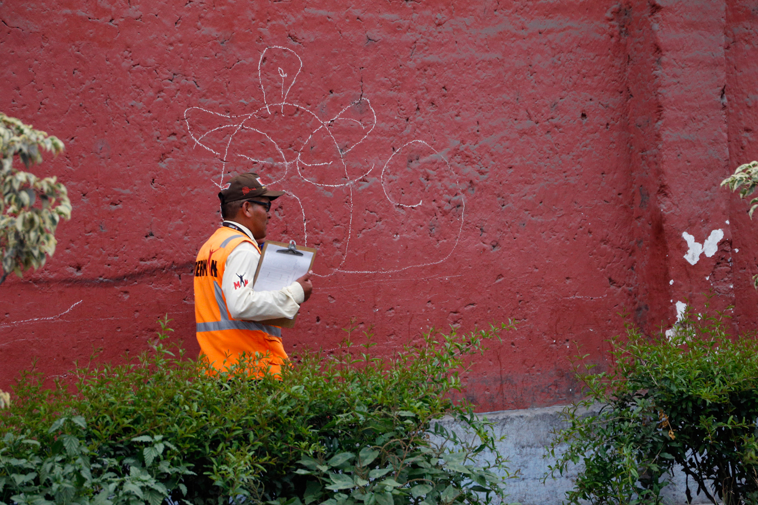 Ein Beamter mit oranger Schutzweste trägt ein Klemmbrett mit einem unbeschriebenen Papierbogen unter dem Arm und läuft an einer rotgestrichenen Mauer vorbei, auf dem ein großes, ungelenkes Graffiti prangt, das ungefähr ein Gesicht darstellt.