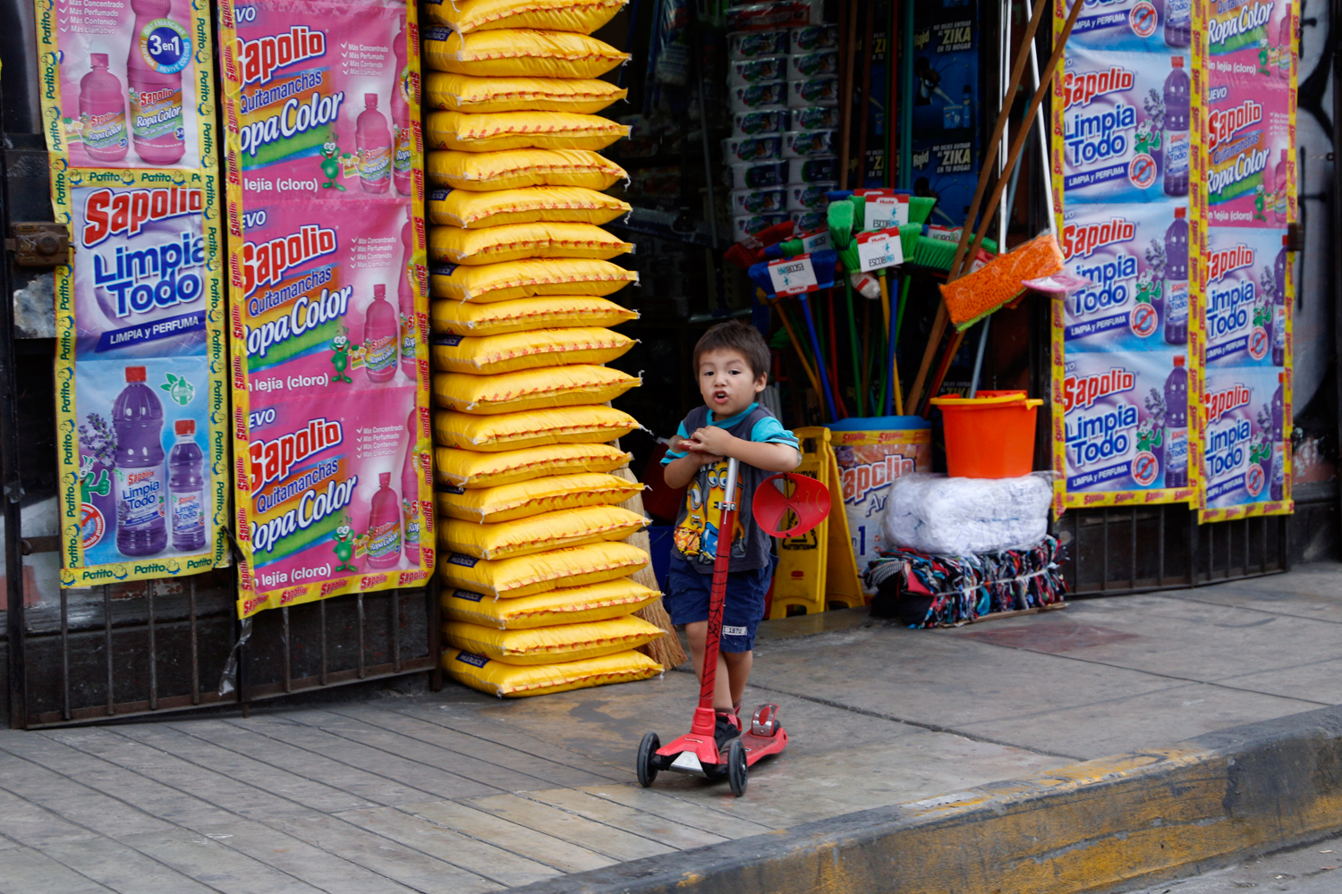 A young boy is riding his red scooter along the pavement in front of a shop that sells cleaning supplies. His shirt features two Minions from the Despicable Me movie franchise and a Spider Man mask is dangling from the handlebar of his scooter.