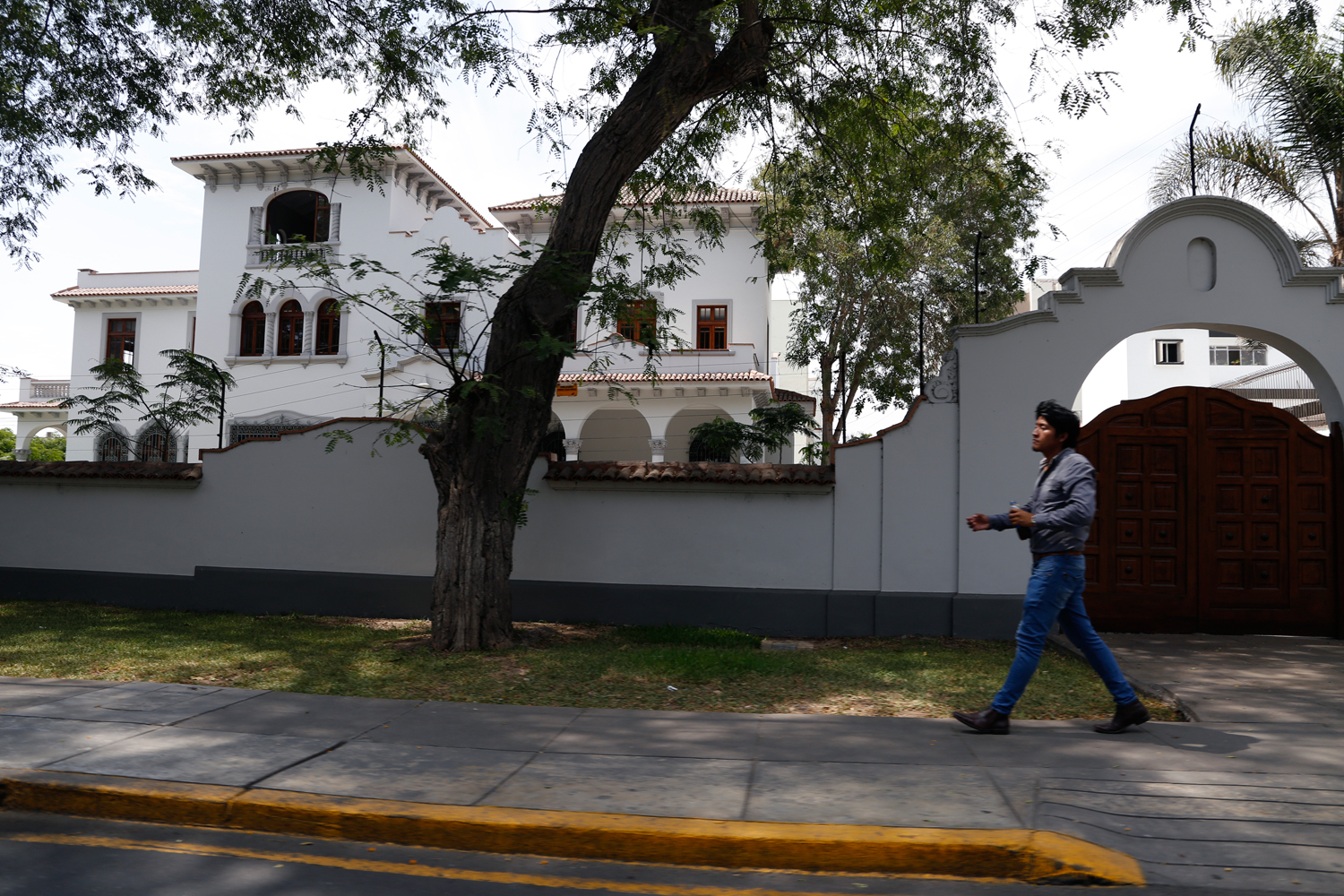 A young man wearing fitted jeans, shiny leather shoes and a dark grey shirt walks along the pavement in an upscale neighbourhood of Lima. He is passing the tall wooden gate to a three-storey, colonial-style villa, painted all white and shaded by a number of trees in its extensive, enclosed garden.