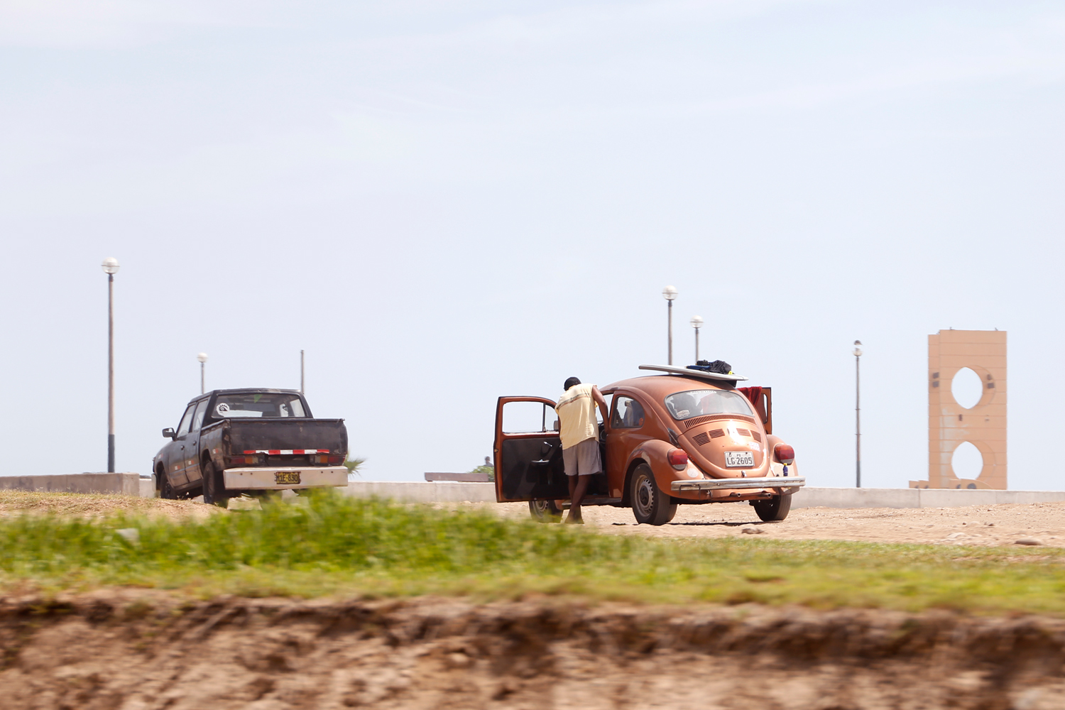 Ein junger Mann lehnt sich in die offene Tür eines bronzefarbenen VW-Käfers, der in der Nähe des Strands geparkt ist. Ein Surfbrett und ein Neoprenanzug liegen auf dem Dach, über der geöffneten Tür auf der Beifahrer*innenseite hängt ein Handtuch.