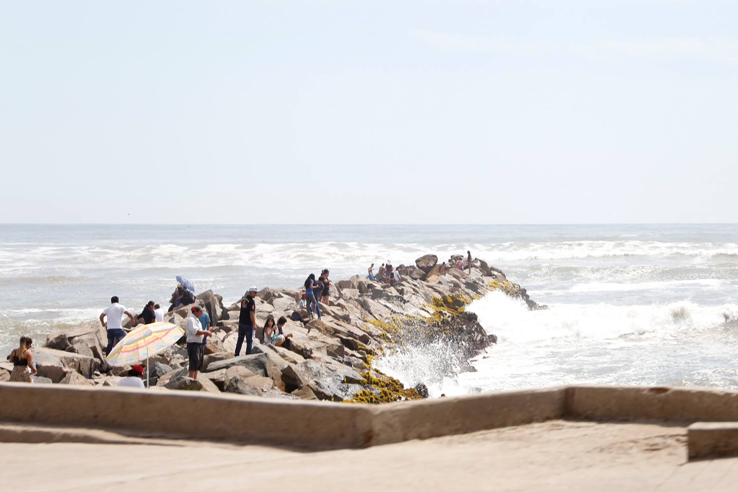 Rund zwei Dutzend Leute sind zu einem felsigen Pier gekommen, der sich von der Strandpromenade ins Meer erstreckt. Manche sitzen auf den Felsen, andere stehen in Grüppchen zusammen, während sich am Rande des Piers die hohen Wellen brechen.