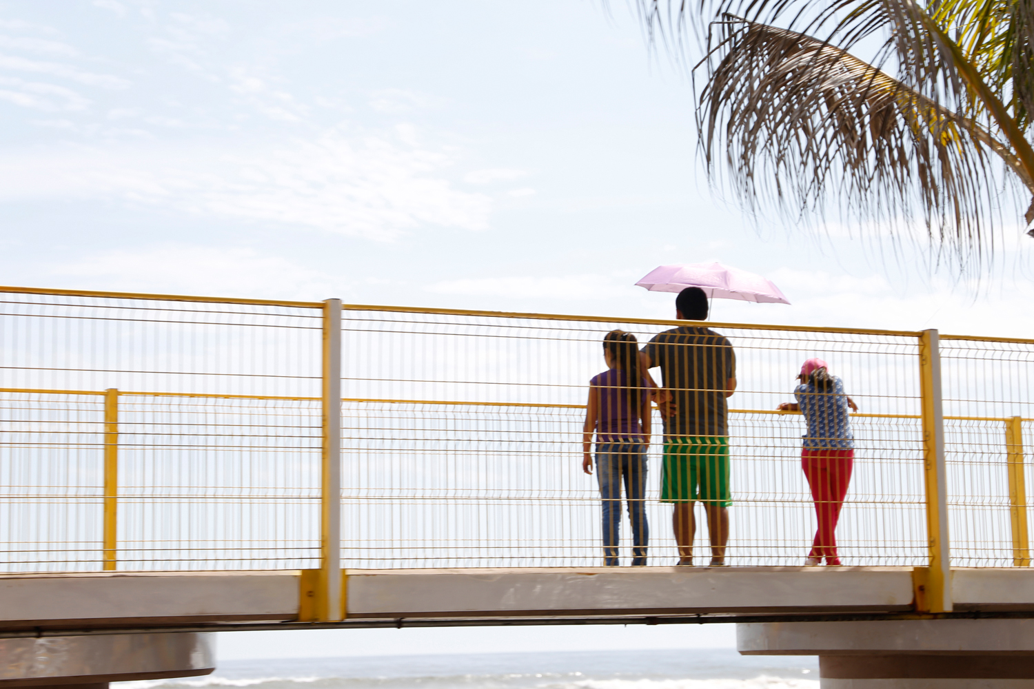 A man and two girls make a stop on a pedestrian bridge overlooking the beach. Shielded from the sun by a pink parasol, they look at the calm ocean underneath a clear blue sky.