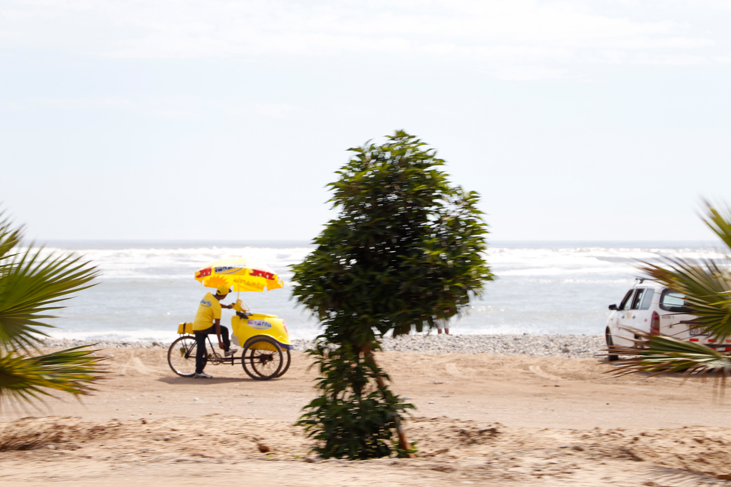 Ein Mann, der Eis der Marke D'Onofrio verkauft, steht neben seinem leuchtend gelben Fahrrad am Strand. Am Lenker ist eine Kühltruhe angebracht und ein großer Sonnenschirm überspannt das gesamte Gefährt.