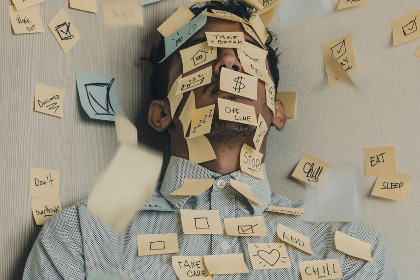 Invoking the expression of somebody drowning in tasks, a neatly dressed man sitting in a chair and with his eyes closed is covered in Post-it notes that are sticking to his face, shirt and the walls behind him. The notes read 'Eat', 'Sleep', 'Work out', 'Take a break', 'Don't overthink', among many more.