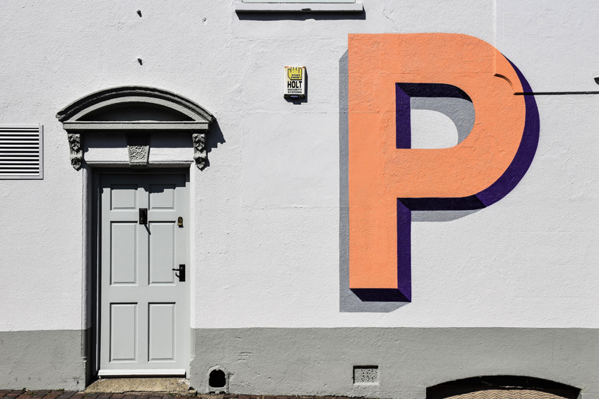 Der leuchtend orange Großbuchstabe P prangt an einer weißen Hausfassade neben einer weißen Tür.