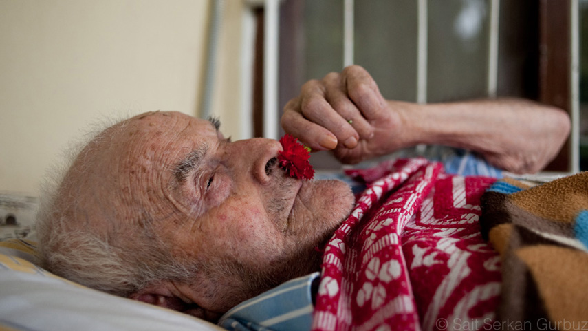 An old Armenian man is lying on the bed and holding a shining red carnation to his nose.